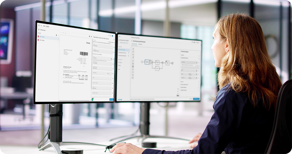 Ansicht Frau vor 2 Bildschirmen mit Workflowdiagrammen und Dokumenten